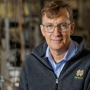 Graham Peaslee, White, is the picture in the gray Quarter Zip Pulovers with a logo of ND Physics in gold on the left side of the chest. It has short, light brown hair and glasses. His laboratory is shown in the background, out of focus.